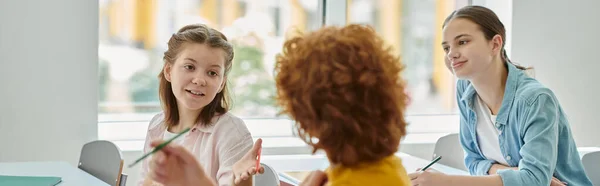 Teen schoolgirl pointing with hand and talking to classmate during lesson in classroom, banner — Stock Photo