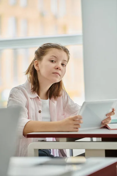 Focused teenage schoolgirl holding digital tablet and looking at camera during lesson in classroom — Stock Photo