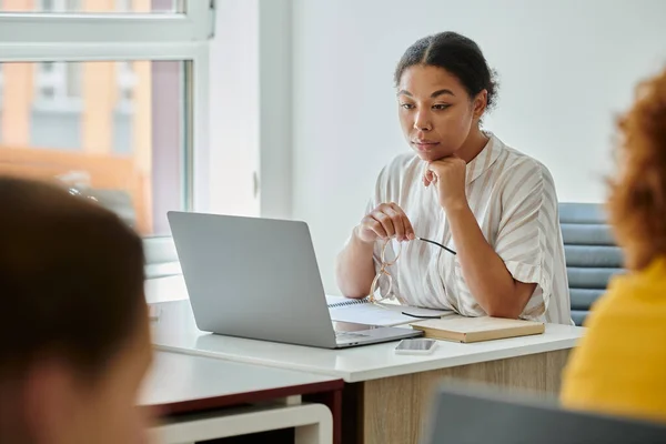 African american teacher holding eyeglasses and looking at laptop near blurred pupils in classroom — Stock Photo