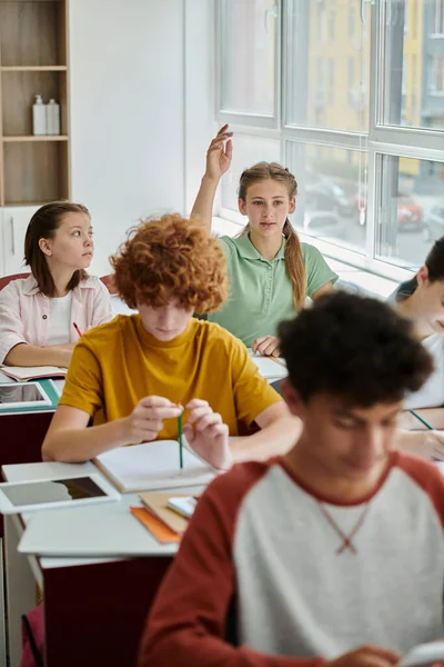 Adolescente escolar levantando la mano y hablando cerca de dispositivos y compañeros de clase durante la lección en la escuela - foto de stock