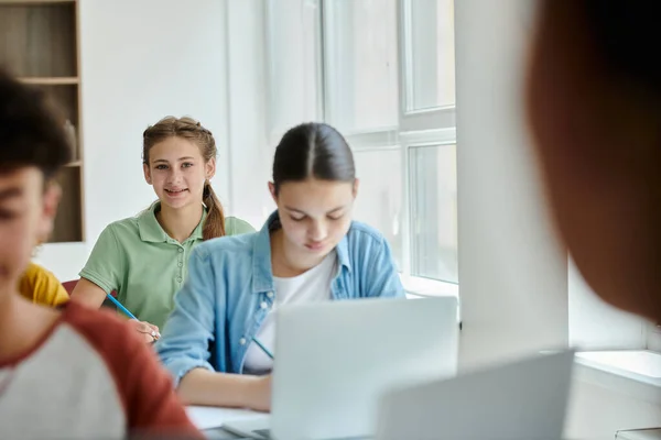 Teen schoolgirl looking at camera and smiling near blurred classmates during lesson in classroom — Stock Photo