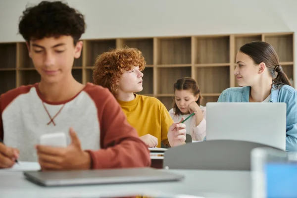 Pelirroja adolescente escolar sosteniendo lápiz y hablando con su compañero de clase cerca de dispositivos durante la lección en clase - foto de stock
