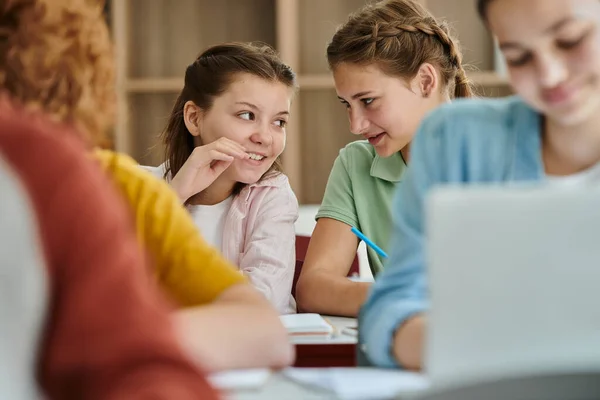 Smiling teenage schoolgirls talking near classmates during lesson in blurred classroom in school — Stock Photo