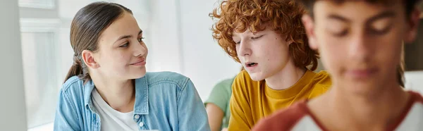 Lächelndes Teenager-Schulmädchen mit Blick auf Klassenkameradin, die während des Unterrichts im Klassenzimmer in der Schule redet, Banner — Stockfoto