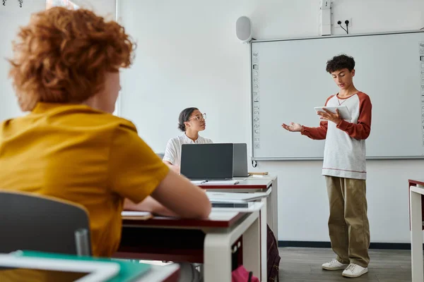 Teenage schoolboy talking while holding digital tablet near african american teacher in classroom — Stock Photo