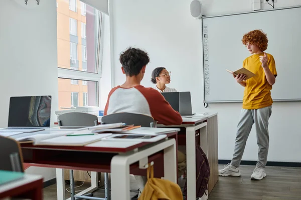 Redhead teen schoolboy holding book and talking near african american teacher during lesson in class — Stock Photo