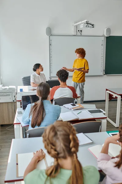 Adolescent écolier tenue livre et parler à afro-américain enseignant pendant leçon dans salle de classe — Photo de stock