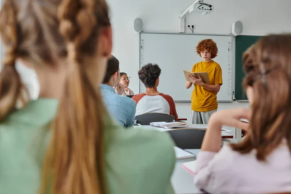 Redhead pupil holding book and talking near african american teacher and friends in classroom — Stock Photo