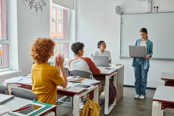 Teen schoolgirl holding laptop during lesson near african american teacher and pupils in classroom — Stock Photo