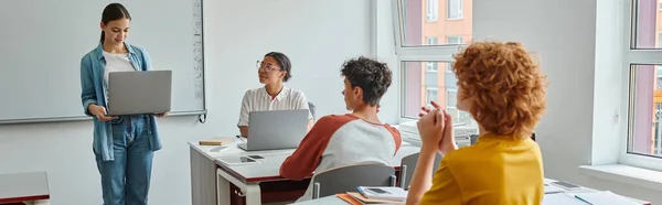 Schoolgirl holding laptop near african american teacher during lesson in classroom, banner — Stock Photo