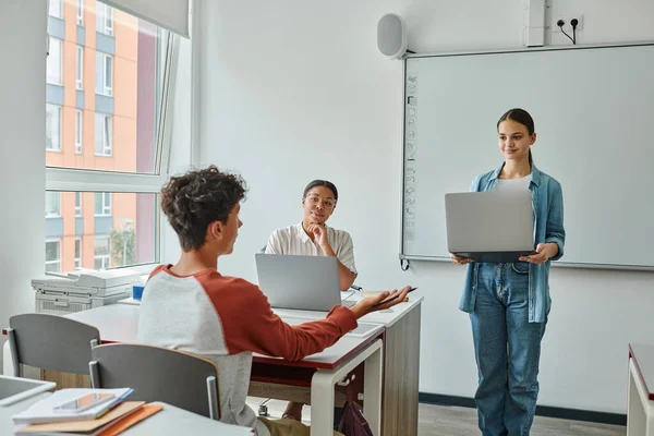 Teen schoolboy talking to classmate with laptop near african american teacher in classroom — Stock Photo