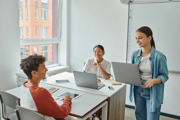 Smiling schoolgirl holding laptop and talking to classmate near african american teacher in class — Stock Photo