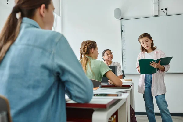 Schoolgirl talking while holding notebook near smiling african american teacher in classroom — Stock Photo
