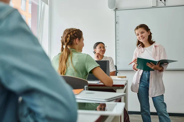 Positive teen schülerin holding notizbuch in der nähe von kamerad und afrikanisch amerikanisch lehrer im klassenzimmer — Stockfoto