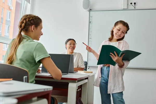 Smiling schoolgirl talking and holding notebook near african american teacher and classmate in class — Stock Photo