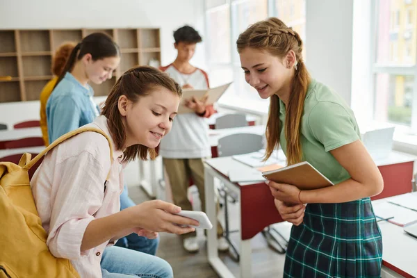 Escolar adolescente sonriente con mochila mostrando smartphone a amigo con portátil en el aula - foto de stock
