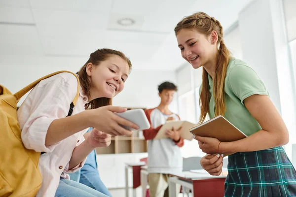 Smiling teen schoolgirls with backpack and notebook using smartphone in classroom in school — Stock Photo