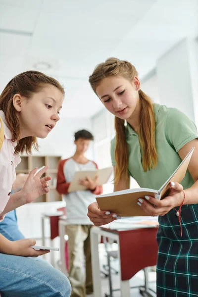 Colegiala adolescente mostrando cuaderno a compañero de clase con teléfono inteligente en el aula borrosa en la escuela - foto de stock