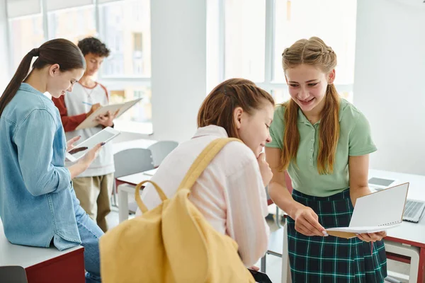 Cheerful schoolgirl holding notebook near friend with smartphone and backpack in classroom — Stock Photo