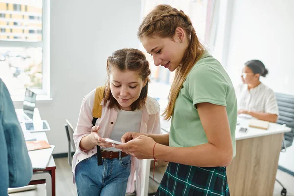 Escolar adolescente sonriente mostrando smartphone a amigo con mochila en aula borrosa en la escuela - foto de stock