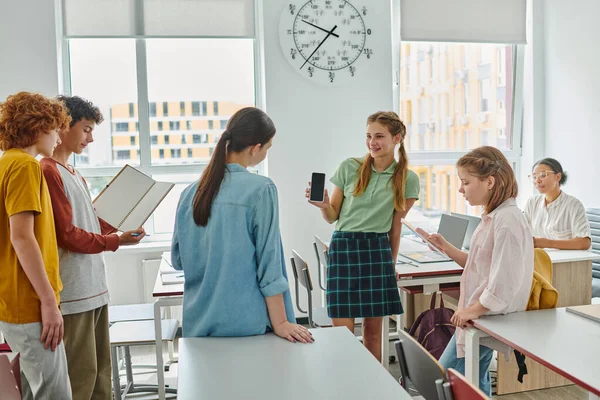 Smiling teen schoolgirl showing smartphone to friends while standing in classroom in school — Stock Photo