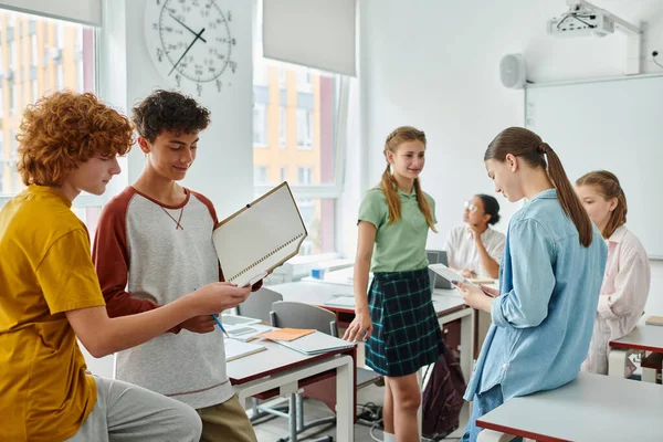 Positive schoolboys holding notebook near friends and african american teacher in classroom — Stock Photo