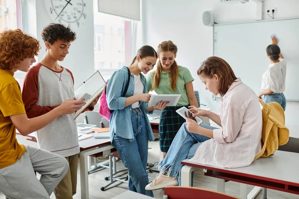 Alunos adolescentes alegres usando dispositivos perto de professor da américa africana em segundo plano em sala de aula — Fotografia de Stock