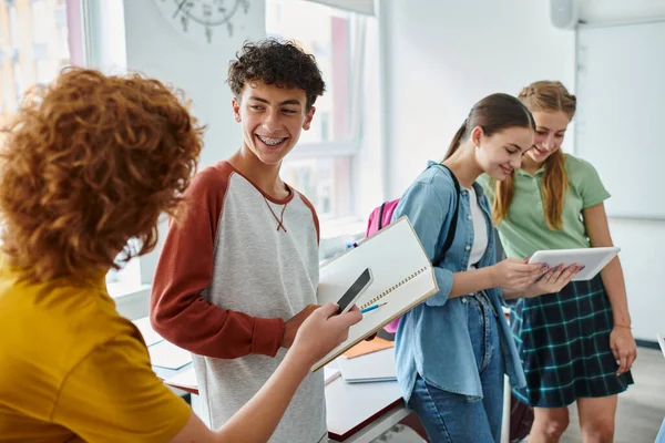 Escolar sonriente con frenos sosteniendo cuaderno cerca de amigos con dispositivos en el aula en la escuela - foto de stock