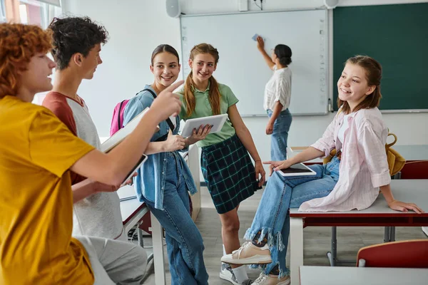 Улыбающиеся школьники разговаривают и указывают пальцем на школьниц с устройствами в классе — стоковое фото