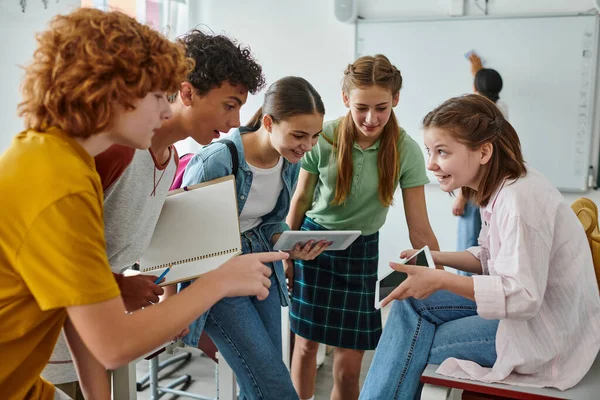 Smiling teen pupils using digital tablet together in classroom near blurred african american teacher — Stock Photo