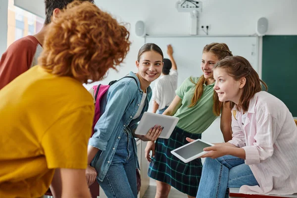 Smiling teen schoolgirls holding digital tablets near blurred friends in classroom in school — Stock Photo