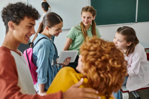 Cheerful teenage pupils talking and using digital tablet near blurred friends in classroom in school — Stock Photo