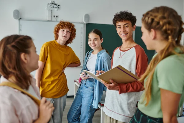 Smiling teenage schoolboy with notebook talking to friends while standing in classroom in school — Stock Photo
