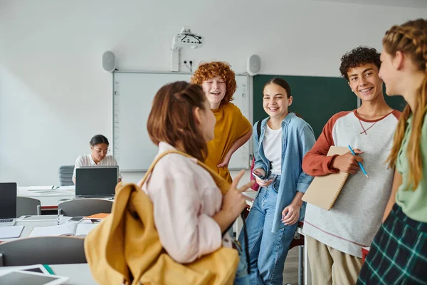 Cheerful teenage pupils with notebook and smartphone standing near blurred friend in classroom — Stock Photo