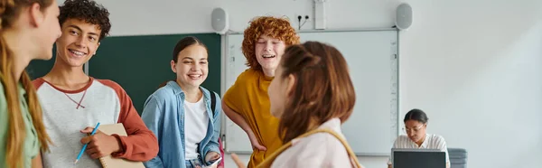 Des camarades de classe adolescents joyeux avec smartphone et ordinateur portable debout près d'un ami dans la salle de classe, bannière — Photo de stock
