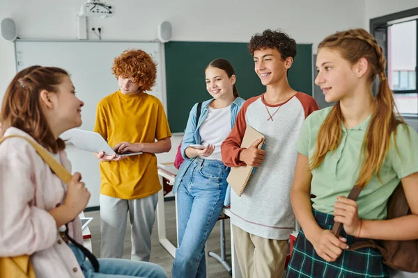 Позитивные подростки с устройствами и рюкзаками разговаривают, стоя в классе в школе — стоковое фото