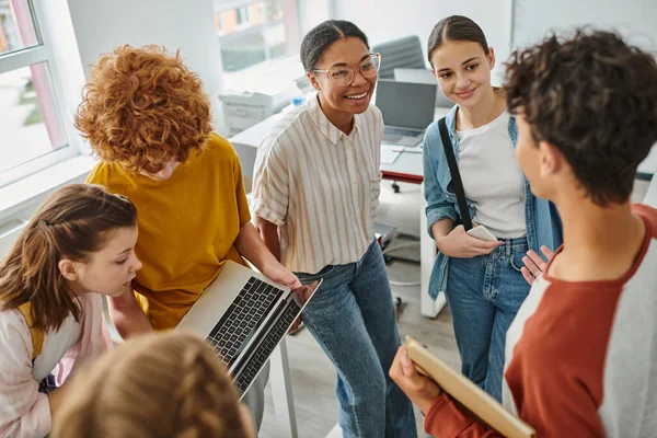 Redhead boy showing laptop to classmate, african american teacher smiling near students — Stock Photo