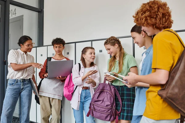 Счастливые студенты-подростки, держащие устройства в школьном коридоре, африканская американка, стоящая рядом с парнем — стоковое фото