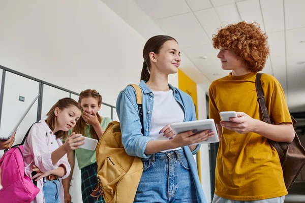 Feliz adolescente y niña sosteniendo gadgets cerca de compañeros de clase en el pasillo de la escuela, volver al concepto de la escuela - foto de stock