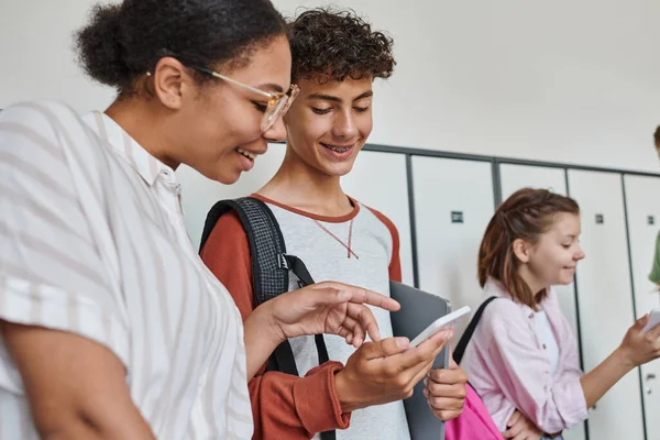 Happy african american teacher pointing at smartphone of student, teenagers in school hallway — Stock Photo