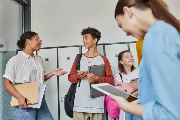 African american teacher gesturing and talking with student, teenagers in school hallway with devices — Stock Photo