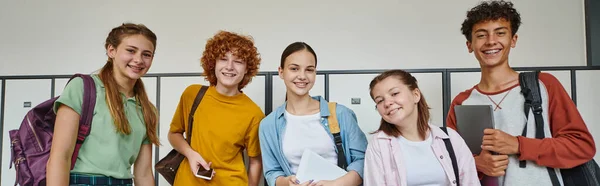 Banner, cheerful teenage students holding devices and looking at camera in school hallway, friends — Stock Photo