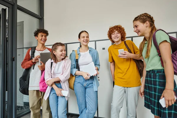 Cheerful teenage students holding devices and chatting in school hallway, teen friends, student life — Stock Photo