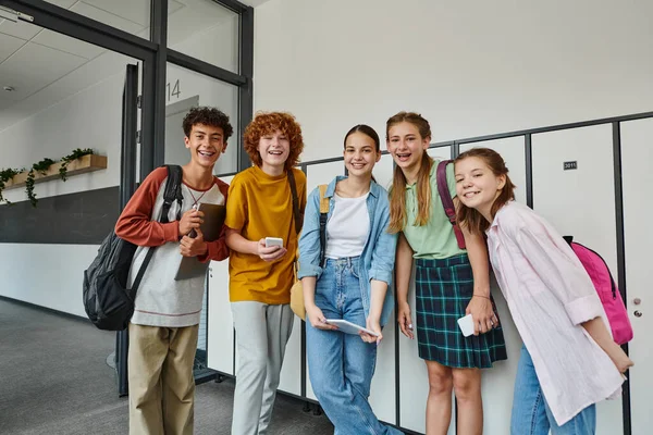 Cheerful teenage students holding devices and looking at camera in school hallway, teen friends — Stock Photo