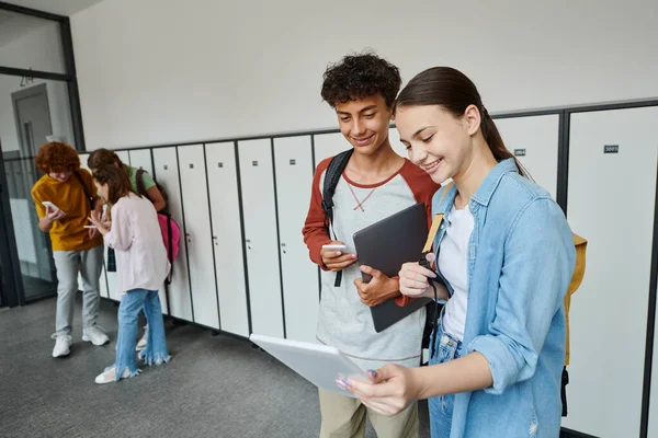 Щасливий хлопчик і дівчинка, дивлячись на цифровий планшет і тримаючи пристрої в шкільному коридорі, підліткові однокласники — стокове фото