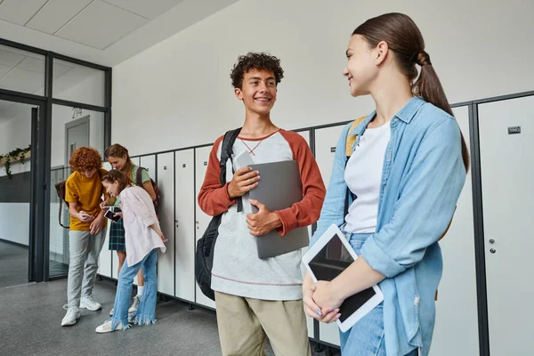 Щасливі школярі тримають пристрої в коридорі, повертаються до школи концепція, посмішка, студенти і технології — стокове фото