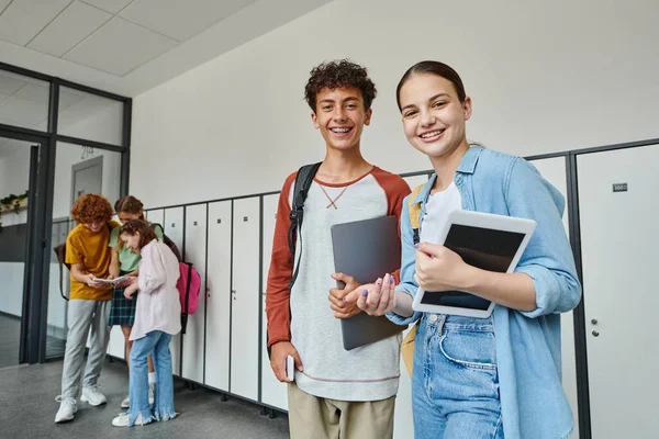 Feliz adolescente compañeros de clase sosteniendo dispositivos y mirando a la cámara en el pasillo de la escuela, estudiantes adolescentes - foto de stock