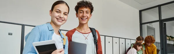 Банер, підліткові однокласники тримають пристрої і дивляться на камеру в шкільному коридорі, щасливі студенти — стокове фото
