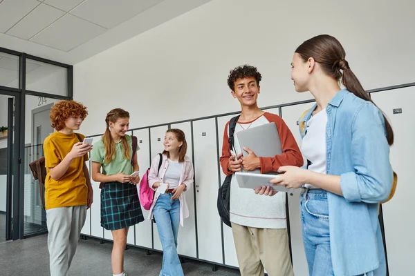Compañeros de clase felices hablando y sosteniendo dispositivos en el pasillo, de vuelta a la escuela, estudiantes adolescentes, era digital - foto de stock