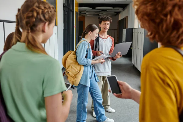 Teen students holding devices and looking at classmates on blurred foreground, school hallway — Stock Photo